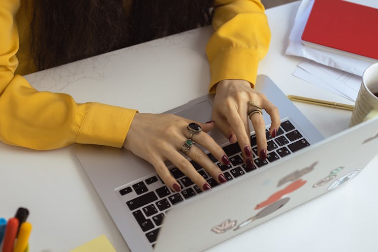 Close-up of a woman typing on a laptop, showcasing red nails and rings, wearing a yellow long sleeve indoors.